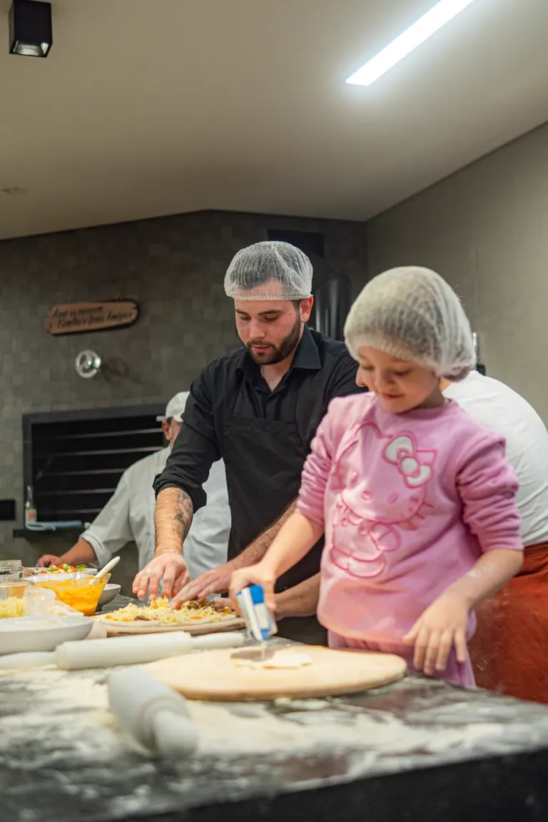 Participantes montando pizzas em bancada com forno ao fundo